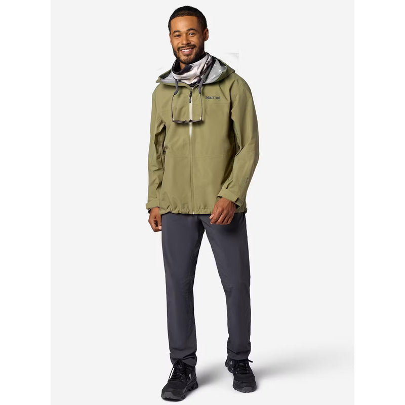 A man modeling an olive green hooded technical jacket with a gray and black patterned neck gaiter, paired with dark gray pants and black shoes, against a light gray studio background.