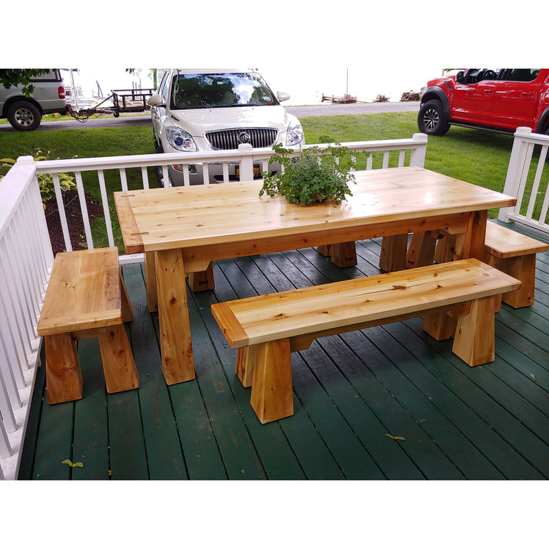 A rustic wooden outdoor dining set consisting of a rectangular table and two matching bench stools, displayed on a green wooden deck with cars visible in the background.