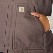 Woman putting her hand in the pocket of a taupe colored vest. A Carhartt logo is visible in the top corner of the pocket.