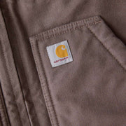 Close up of one of the pockets of a taupe colored vest with a Carhartt logo at the top of it. 