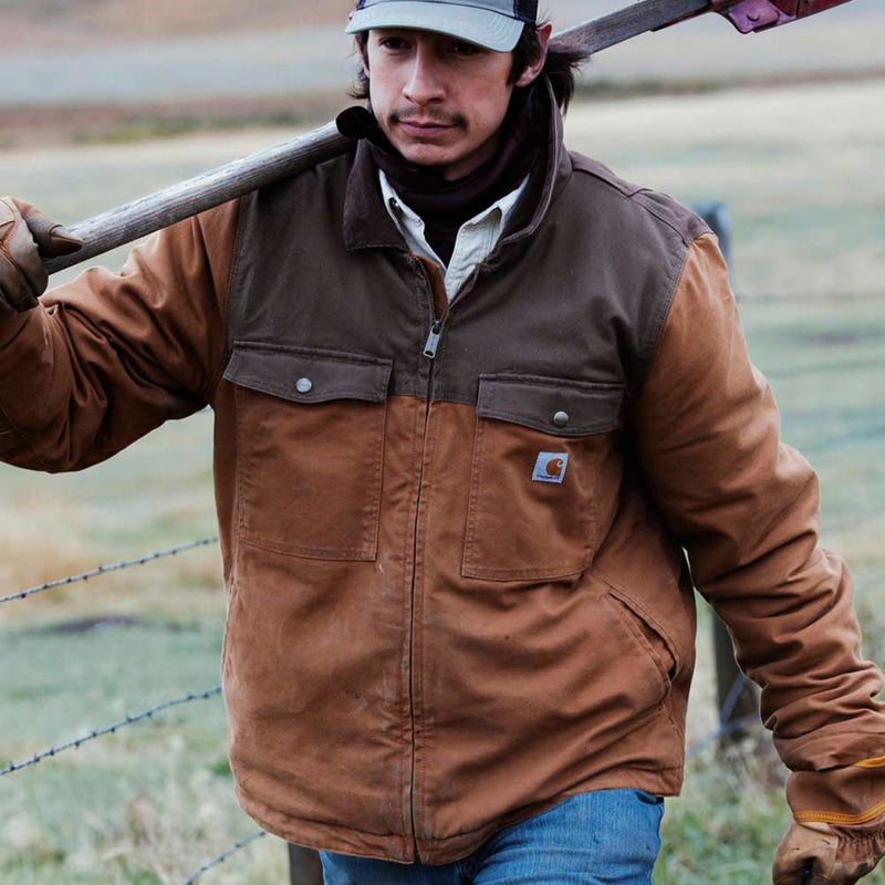 Man carrying a shovel in a field near a fence wearing a brown colored duck-lined insulated jacket. The jacket has two zippered pockets on either side of the chest, as well as a Carhartt logo on the right side pocket.