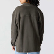 A person shown from the back wearing an oversized waffle-knit button-up shirt in dark olive gray, paired with light blue jeans, against a plain light gray background.