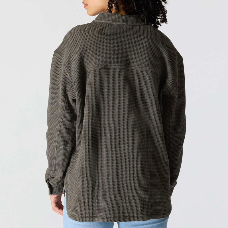 A person shown from the back wearing an oversized waffle-knit button-up shirt in dark olive gray, paired with light blue jeans, against a plain light gray background.