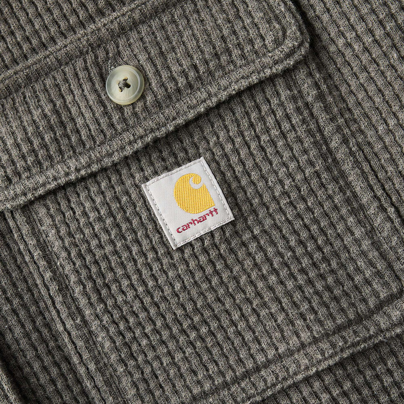 Close-up of a gray ribbed knit jacket or sweater showing a flap pocket with a metal button and a white Carhartt brand patch on the front.