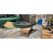 A green and beige canoe resting on a wooden stand inside a workshop or garage, with other boats and equipment visible in the background.