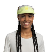 A smiling woman with long black braids wears a light green and white cap with a small yellow square logo patch on the front, paired with a white long-sleeve top against a plain white background.