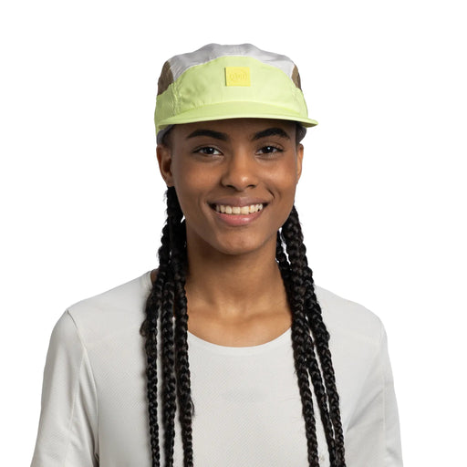 A smiling woman with long black braids wears a light green and white cap with a small yellow square logo patch on the front, paired with a white long-sleeve top against a plain white background.