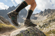 Person standing on a rock wearing grey hiking shoes and black socks with mountains in the background.