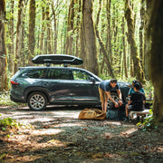 People packing up their bags next to a car in the woods with a black cargo roof rack attached to the top of it.