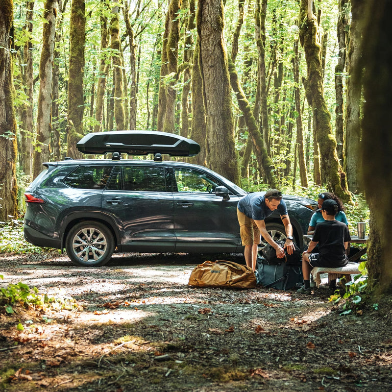 People packing up their bags next to a car in the woods with a black cargo roof rack attached to the top of it.