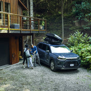 Kid handing a man a skateboard in the woods next to a cabin. The man is putting the skateboard into a black cargo roof rack that is attached to a car. 