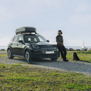 A man and his dog leaning up against a car with a black cargo roof rack attached to it as a boat sails off in the background. 