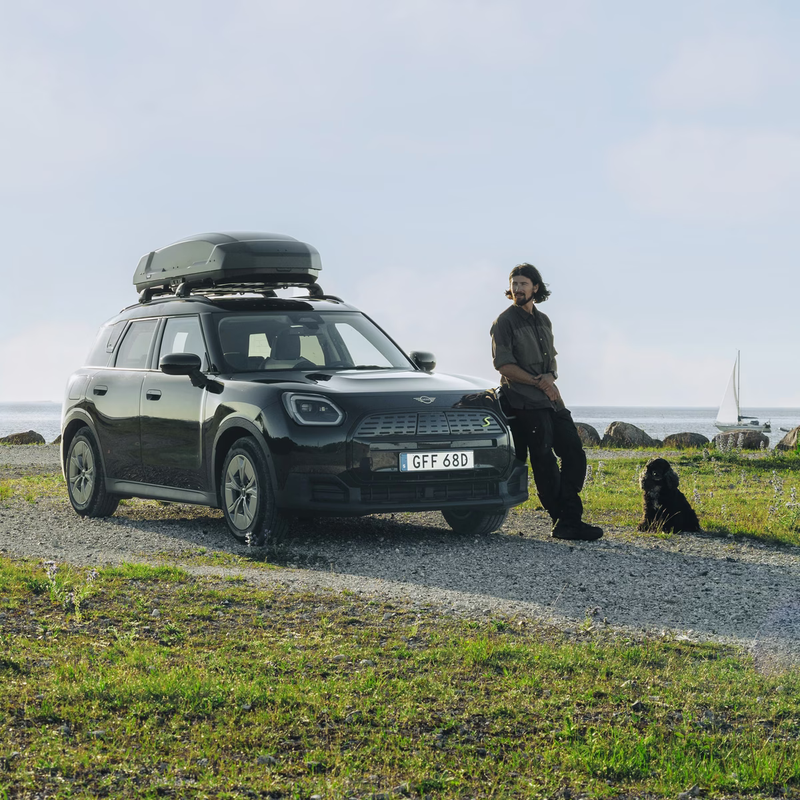 Person standing next to a car with a roof box on a coastal road
