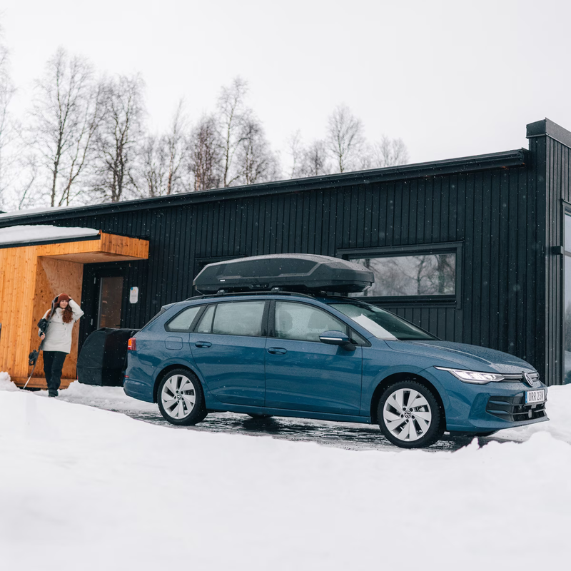 Blue car with a roof box parked in front of a black building in a snowy landscape.