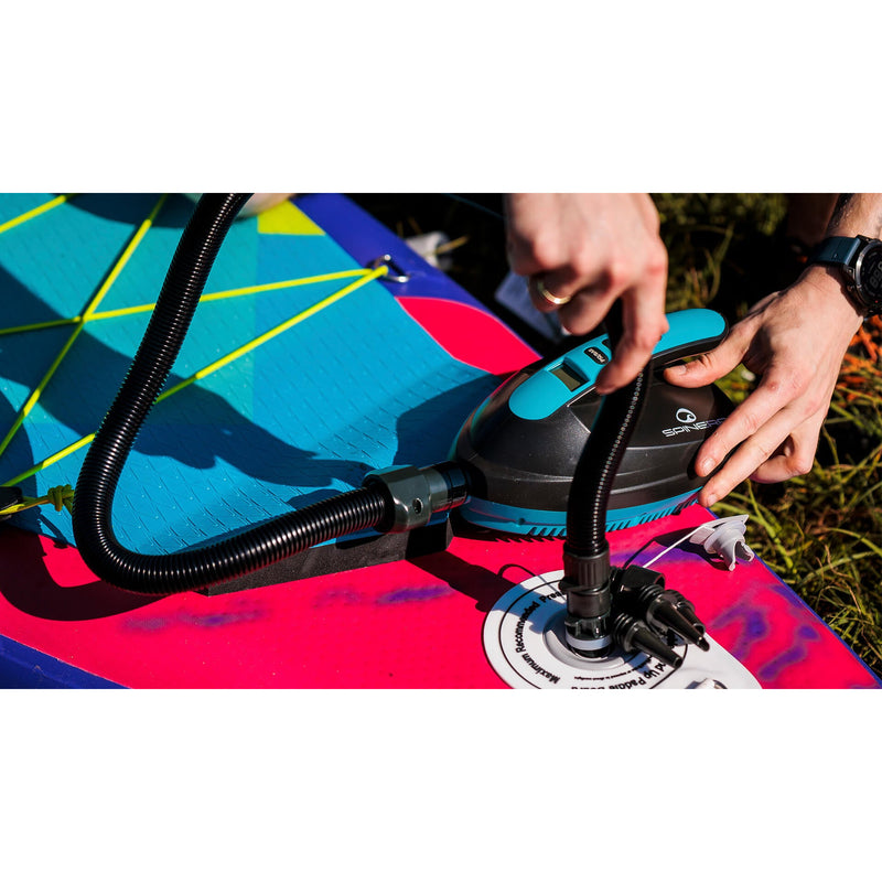 A person using a black and teal pump to inflate a bright pink and blue paddle board outdoors on grass.