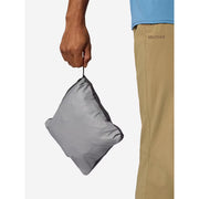 A person holds a compact, folded gray Marmot pouch or bag against a light gray background, with the person's hand, arm, and khaki pants partially visible. The Marmot brand name is printed on the pants.