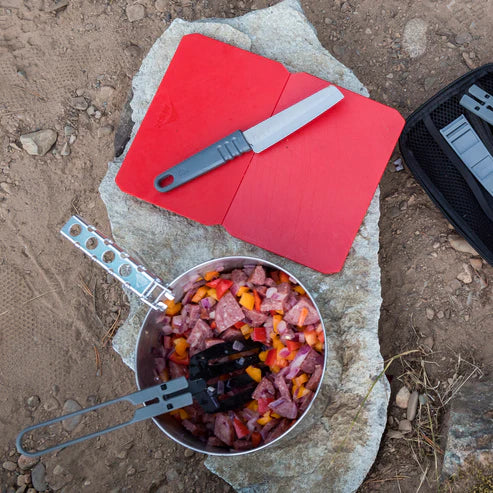 Cooking setup with a pot of food, knife, and cutting boards on a rock outdoors.