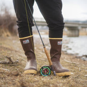 Person wearing brown rubber boots with yellow soles, standing near a river with a fishing rod.