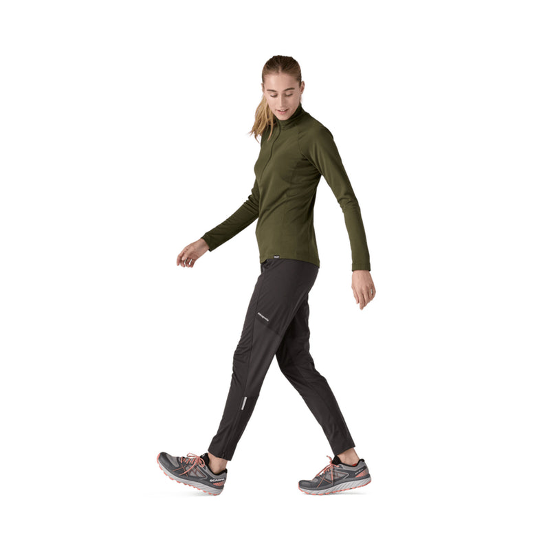 A woman walking in athletic wear, featuring an olive green long-sleeve mock neck top and black running pants, against a solid white background.