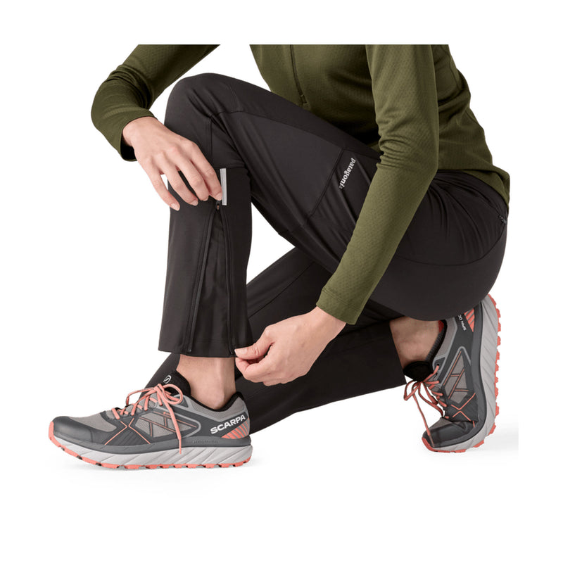 A model wearing black Scarpa hiking pants and gray trail running shoes, crouching down to demonstrate a zippered ankle pocket on the pants against a white background.