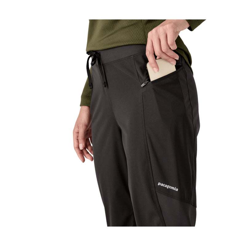A close-up of a person wearing black Patagonia pants, showing a side pocket with a beige phone inserted and the Patagonia logo near the lower leg.