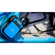 A close-up of a blue and black Spinera electric pump mounted on a bright blue inflatable board, with a hose connected and a blurred outdoor background.