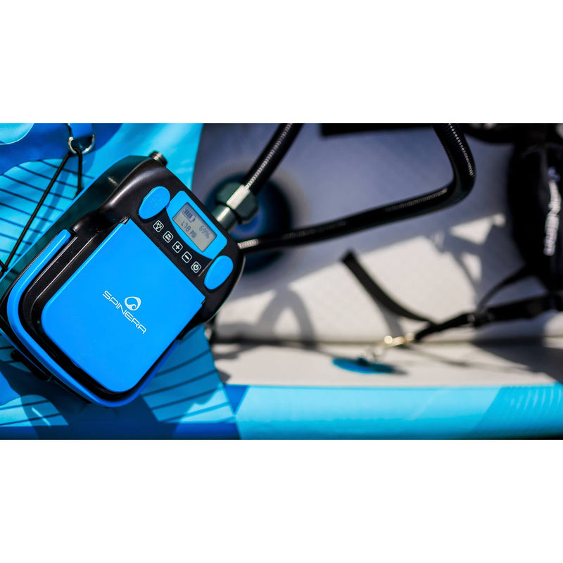 A close-up of a blue and black Spinera electric pump mounted on a bright blue inflatable board, with a hose connected and a blurred outdoor background.
