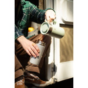 A person pours hot coffee from a mint green handled pitcher into a white insulated travel mug. The person is wearing a green plaid flannel shirt and brown pants, and the scene is shot in warm, natural light next to a white vehicle or trailer door.