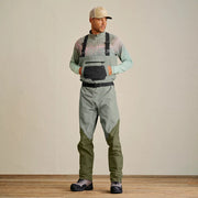 Man looking to the side with his hands in the pockets of his sage colored waders against a gray wall. 