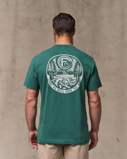 Man wearing a green t-shirt with a logo and text on the back, standing against a beige wall.