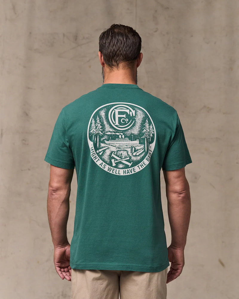 Man wearing a green t-shirt with a logo and text on the back, standing against a beige wall.