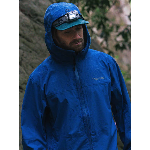 A man wearing a bright blue Marmot hooded rain jacket, a teal cap with a headlamp, and a beard, standing outdoors against a blurred natural background.