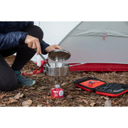 Person preparing a camping meal with a portable stove and cookware near a tent.