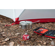 A camping setup on a forest floor with dry leaves, featuring a small stainless steel pot on a red portable gas stove, an open red and black tool case with utensils, and part of a white and red tent in the background.