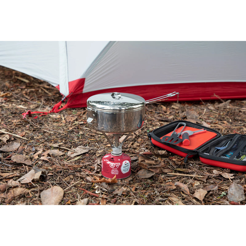 A camping setup on a forest floor with dry leaves, featuring a small stainless steel pot on a red portable gas stove, an open red and black tool case with utensils, and part of a white and red tent in the background.