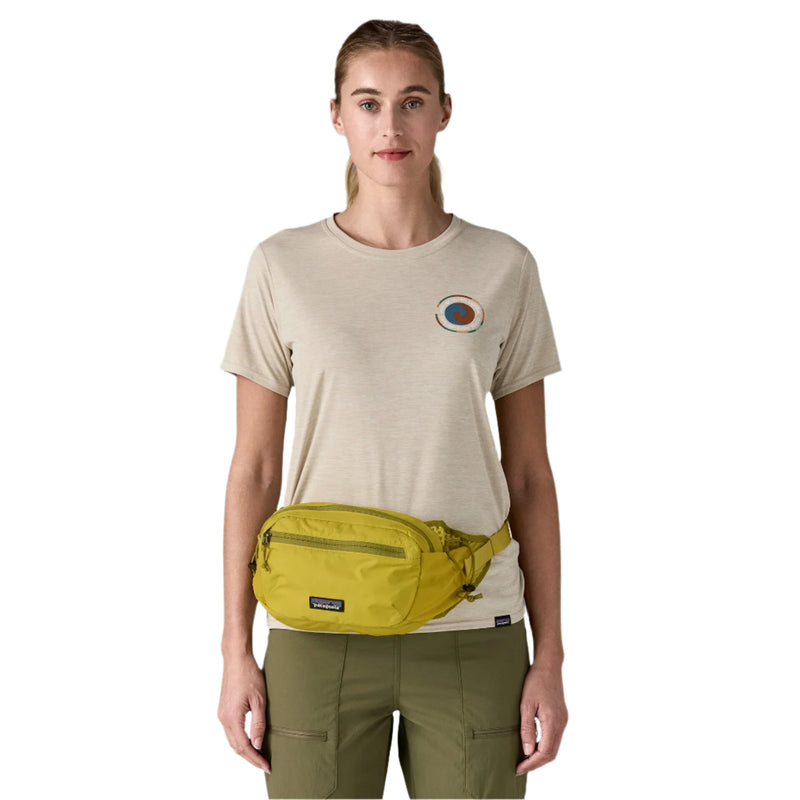 Person wearing a beige t-shirt with a logo and green pants, carrying a yellow waist bag on a white background