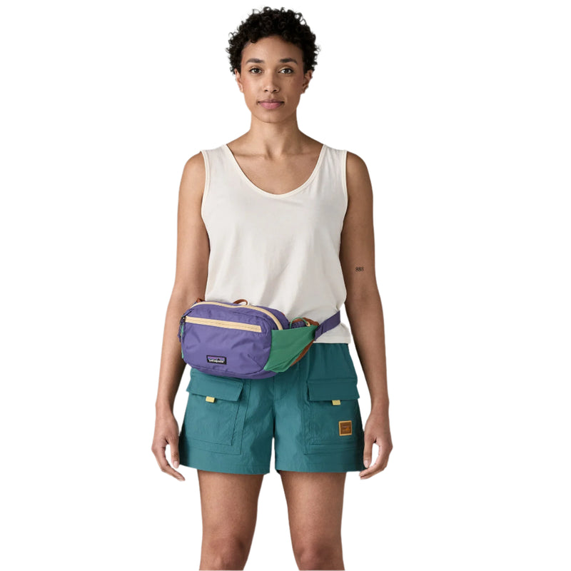 Woman wearing a white tank top and teal shorts with a purple and green fanny pack on a white background
