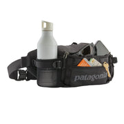 A black waist bag with a water bottle, glasses, phone, keys, cards, and a snack inside on a white background
