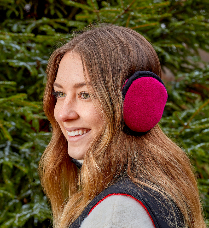 Woman wearing a pair of magenta colored earmuffs against some foliage. 