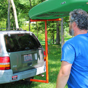 Man loading a green kayak onto a red car carrier attached to a Jeep in a grassy area.