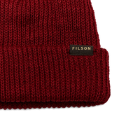 Close up of a red beanie with a tan Filson logo on it against a white background. 