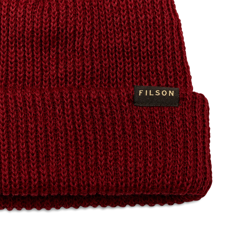Close up of a red beanie with a tan Filson logo on it against a white background. 