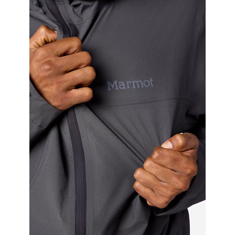 Close-up of a person wearing a dark gray Marmot jacket, pulling on the front  with both hands to demonstrate the stretch. The Marmot logo is visible on the chest.