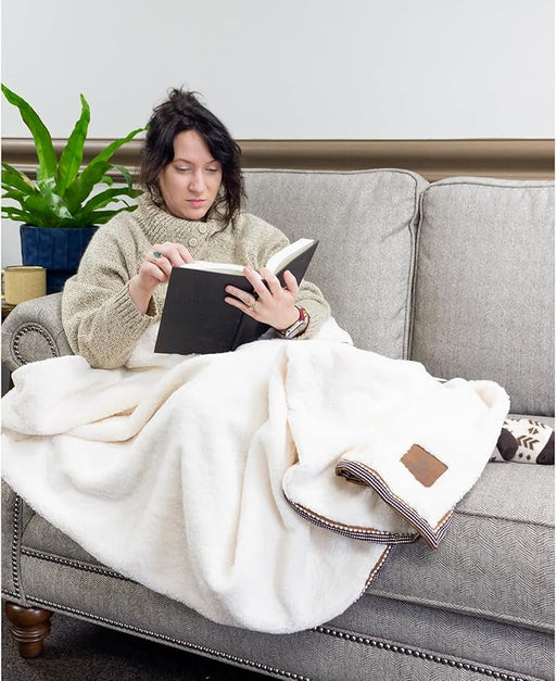 Woman sitting on a couch under a white throw blanket reading a book. 