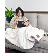 Woman sitting on a couch under a white throw blanket reading a book. 