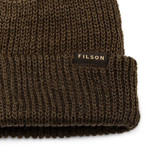 Close up of an olive colored beanie with a tan Filson logo against a white background. 