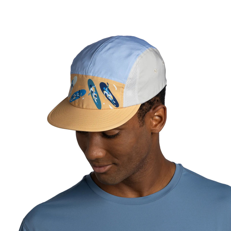 A man wearing a color-blocked 5-panel trucker cap with a light blue front panel, white mesh back panels, and a tan brim with blue surfboard graphics, set against a plain white background.