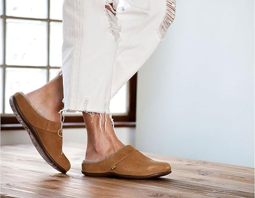 Brown clogs worn with white pants on a wooden floor.