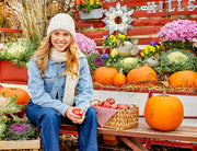 Woman wearing a white knit beanie with brown accents sitting next to some pumpkins. 