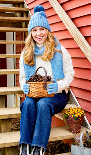 Woman wearing a sky blue knit pom pom beanie holding a basket while sitting on some steps.